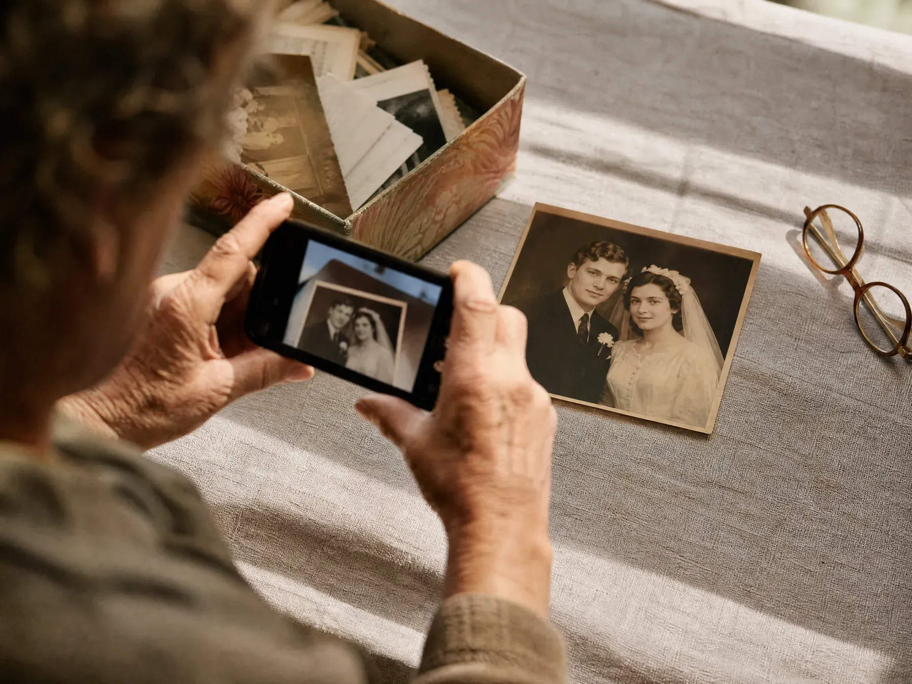 Weathered hands photograph an old wedding print resting on a linen tablecloth beside a box of loose photographs.