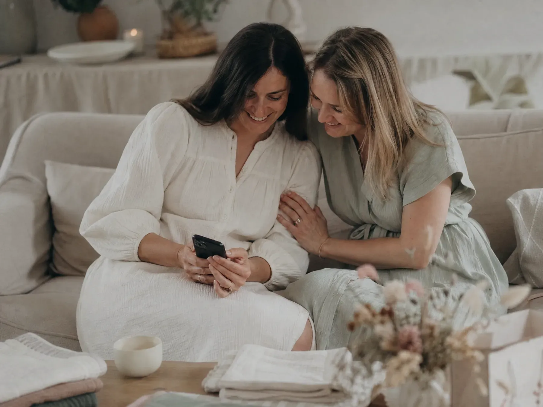 Two women sit close on a sofa writing a message together on a phone at a baby shower.