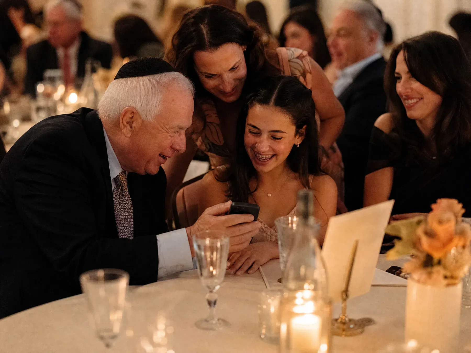 A grandfather in a kippah shows his phone to a teenage girl at a bat mitzvah table while an aunt looks on.