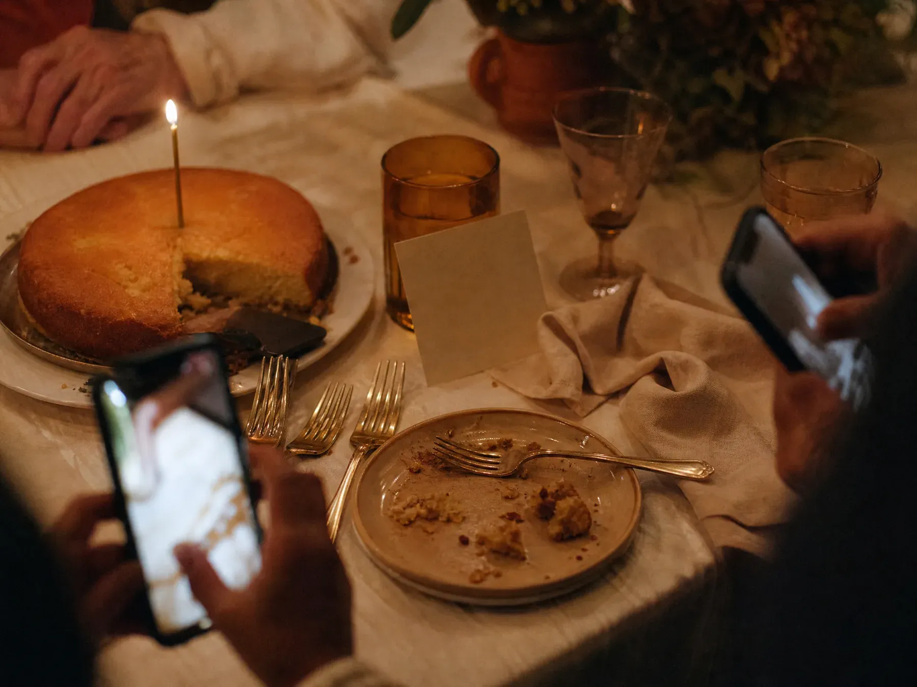A birthday cake with a lit candle beside a small propped card and two hands holding phones over the table.