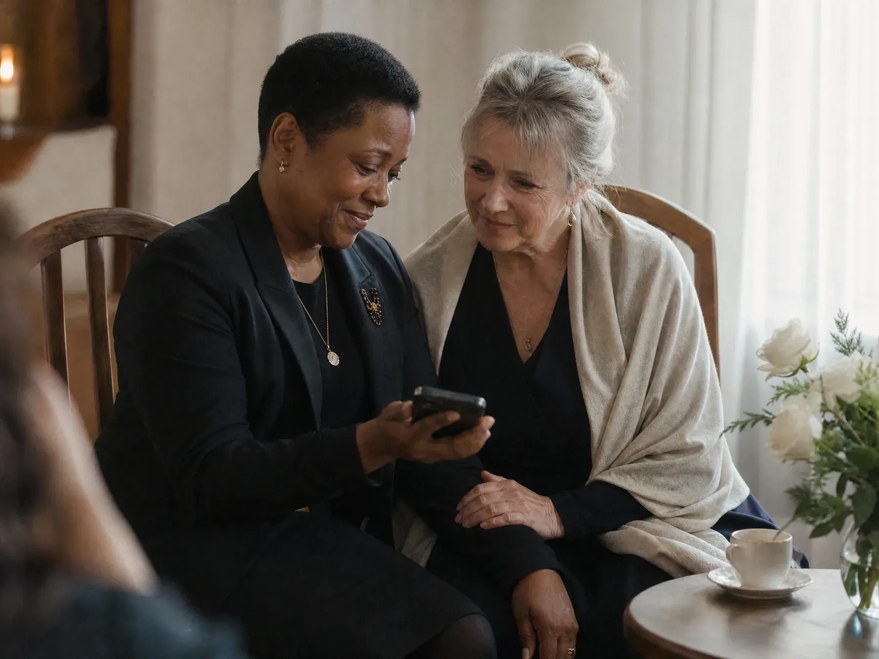 Two people sit side by side at a memorial gathering, one showing the other a photograph on a phone with a hand on their arm.