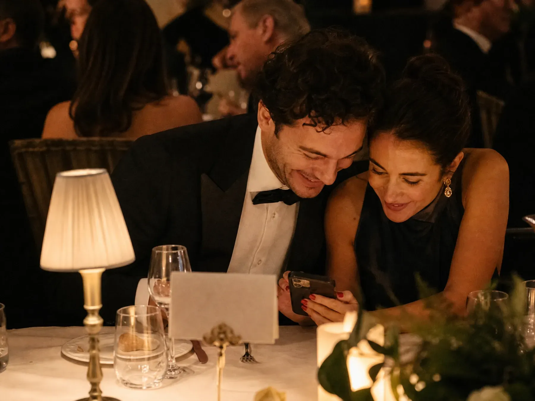 A couple in black tie lean together over a phone at a candlelit gala table beside a small propped card.