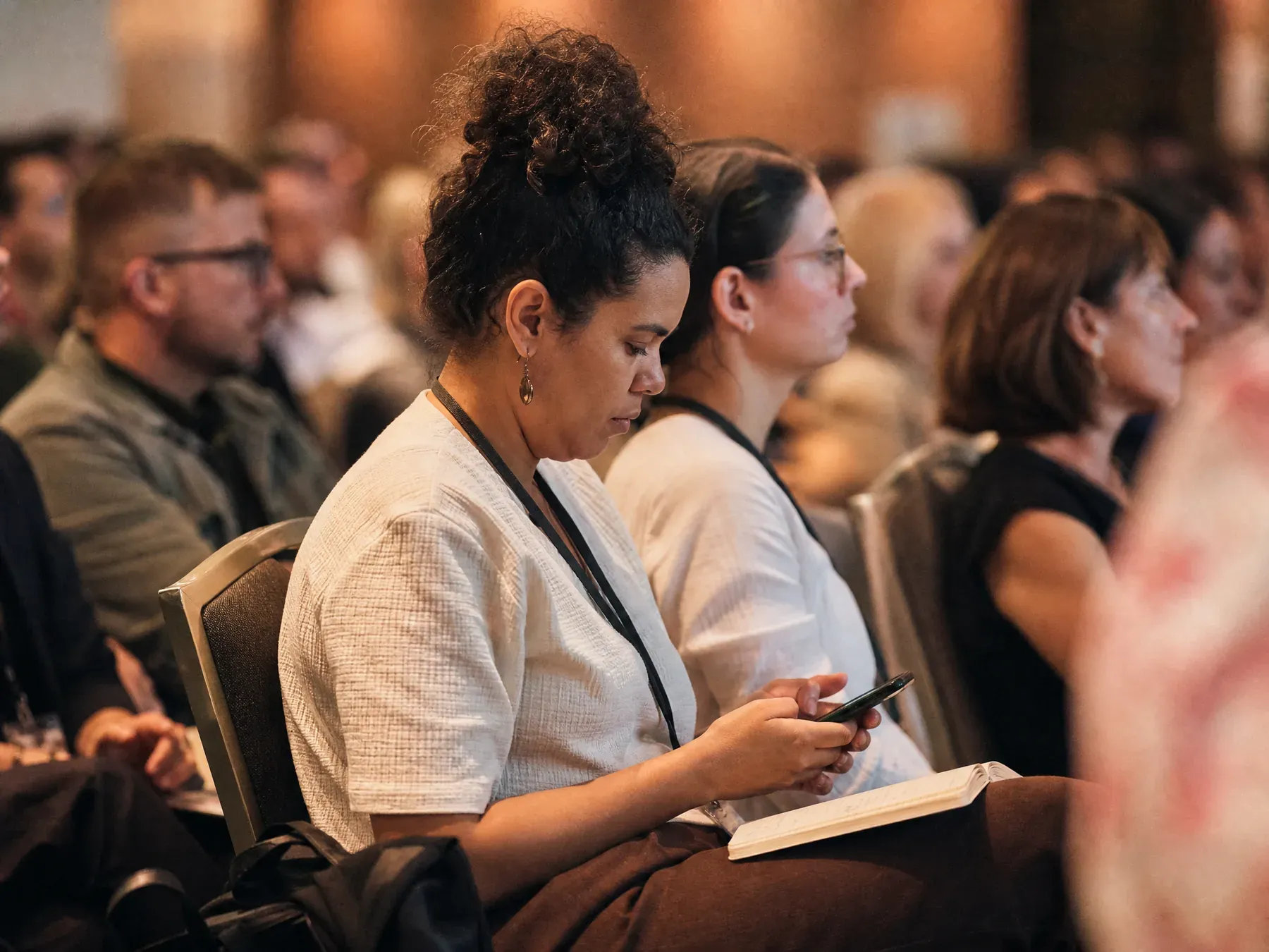 A conference delegate types on her phone mid-row with an open notebook on her knee.