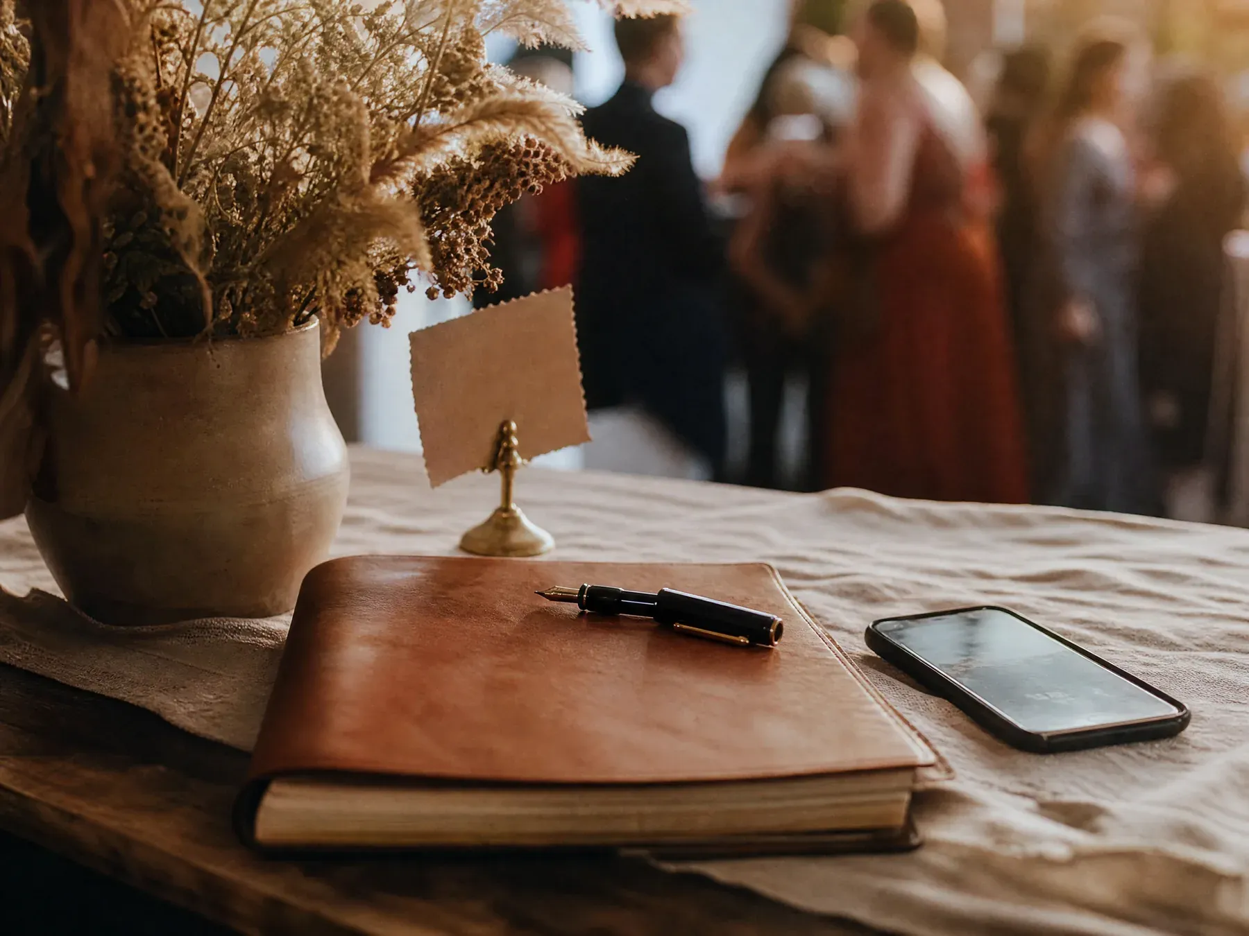 Still life of a closed leather guest book with a fountain pen beside a small propped card and a phone lying face up.