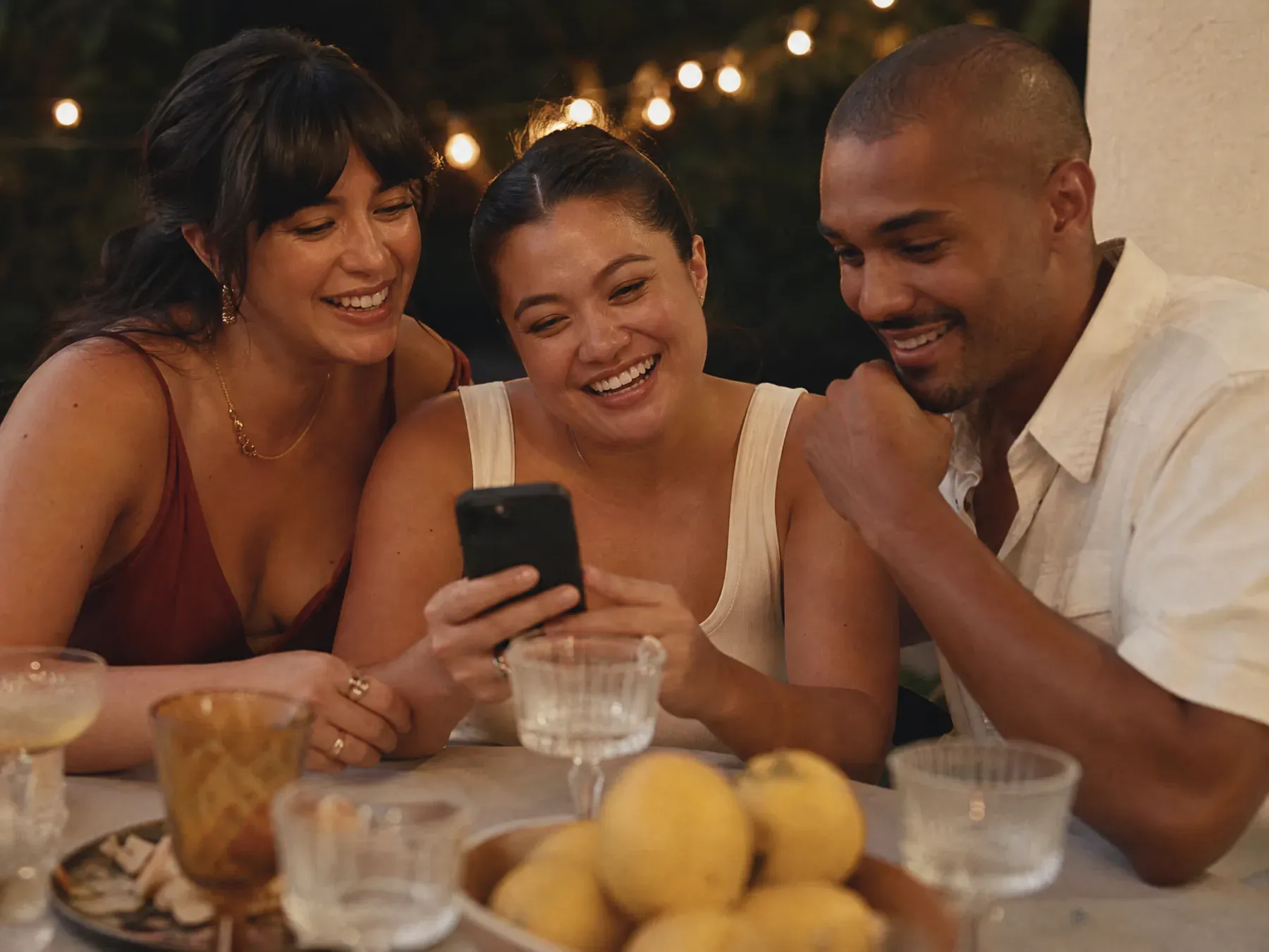 Three friends crowded together on a garden bench composing a message on one phone at an engagement party.