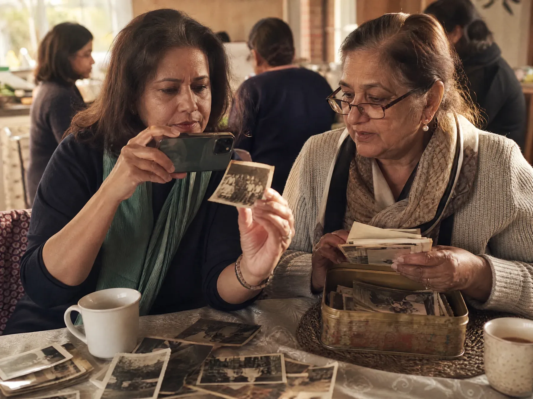 A woman photographs an old curled family print with her phone at a kitchen table while an aunt holds more photographs.