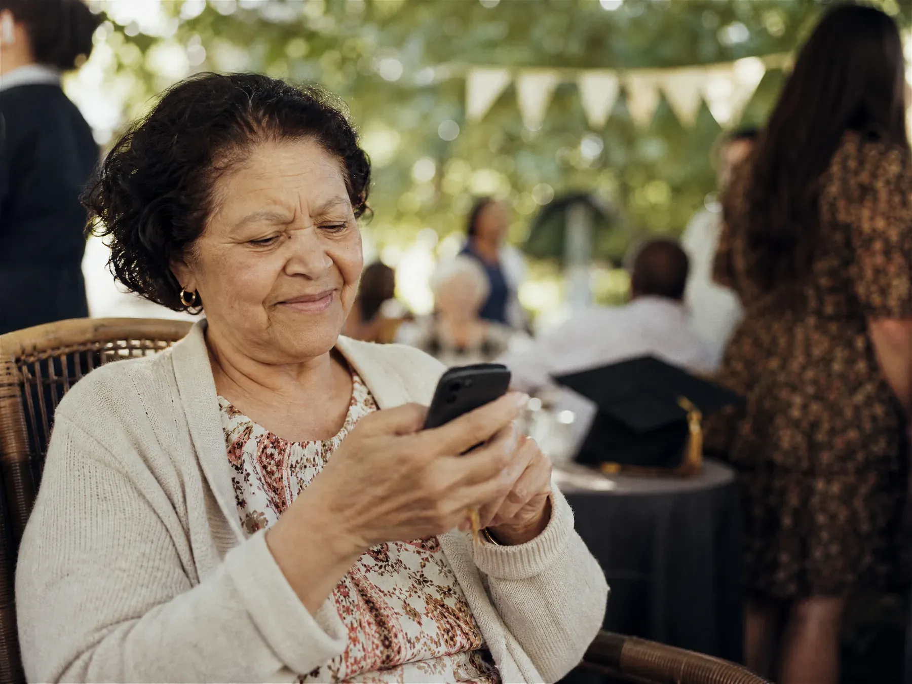 A grandmother concentrates on typing a message on her phone in a garden chair at a graduation party.
