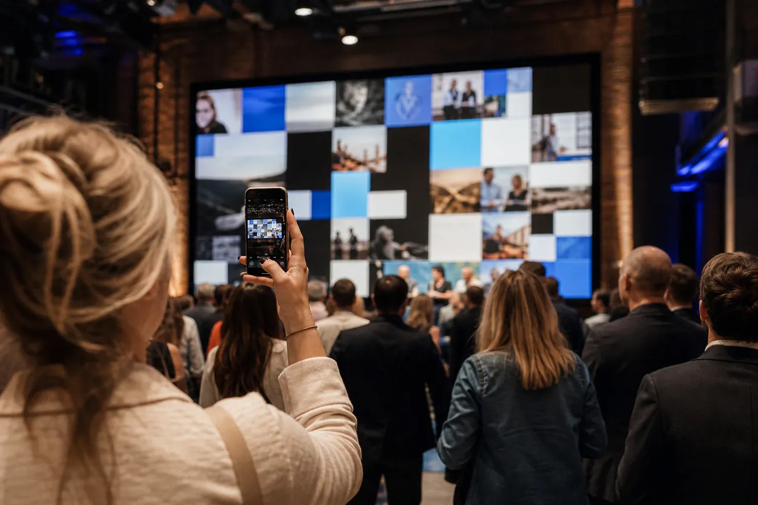 Event attendees beneath a large live wall as one person holds a phone showing a social post composer.