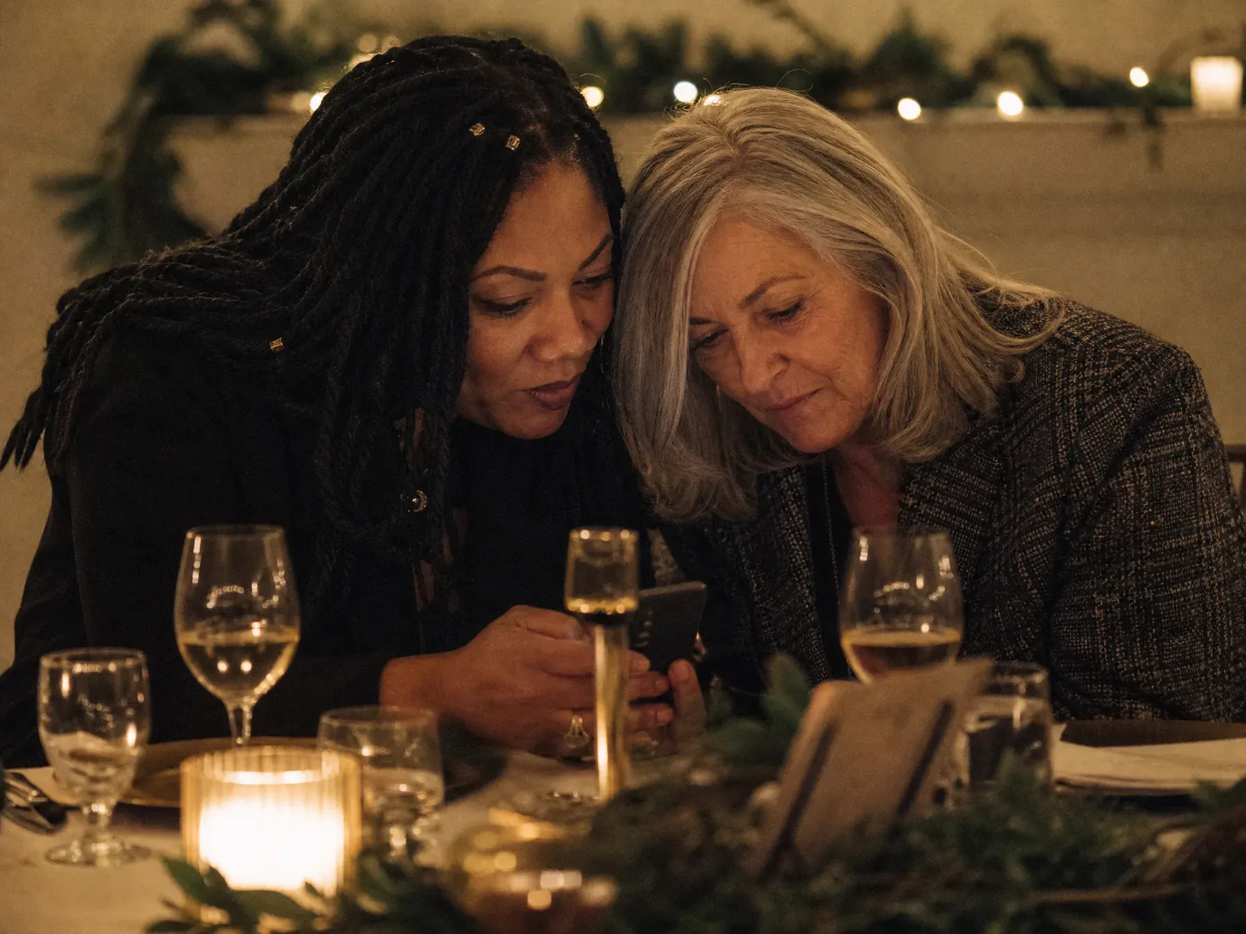 Two colleagues lean together over a phone by candlelight at a holiday party table with a sprig of pine beside them.