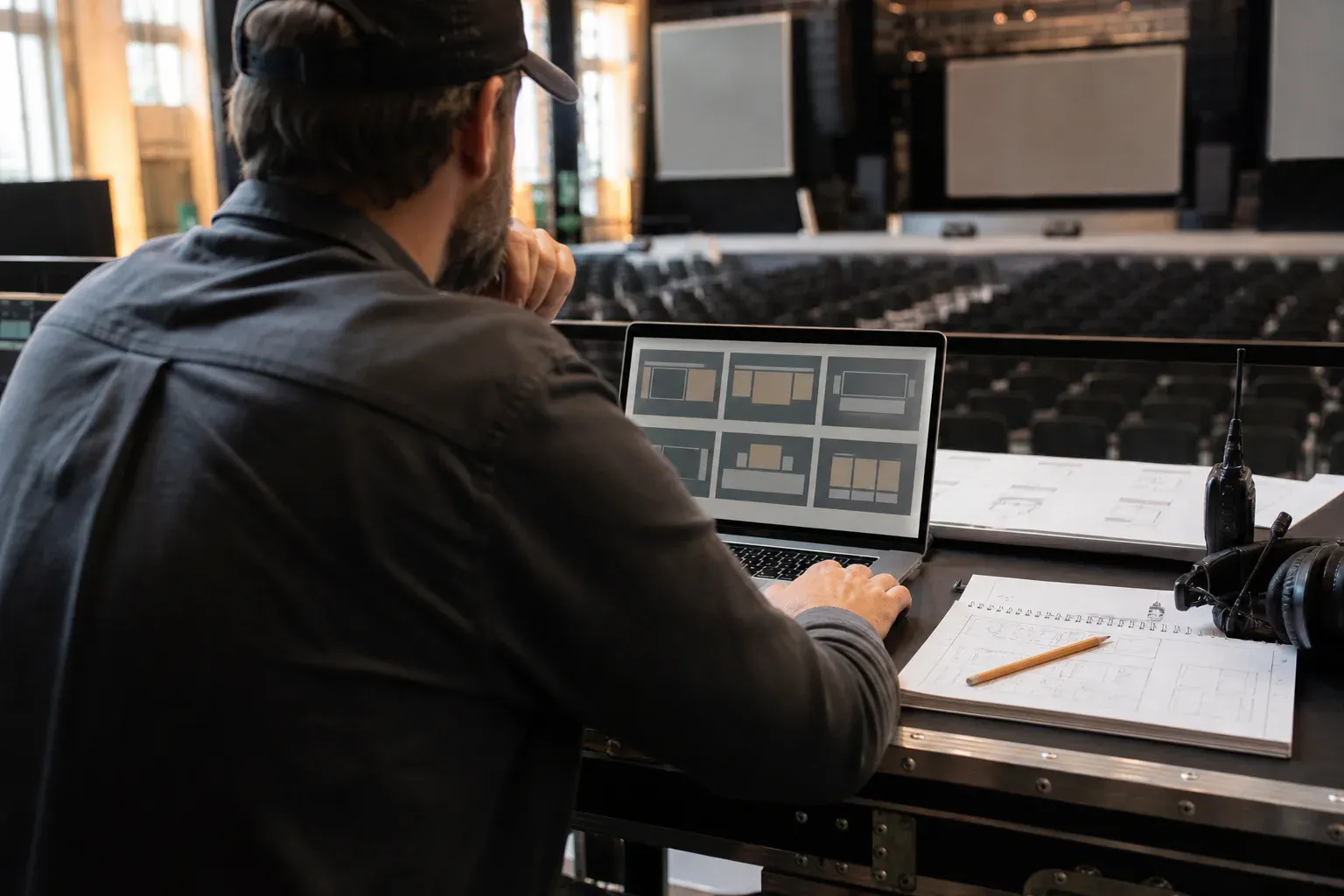An event producer comparing live wall layouts on a laptop beside a production run sheet and venue radio.