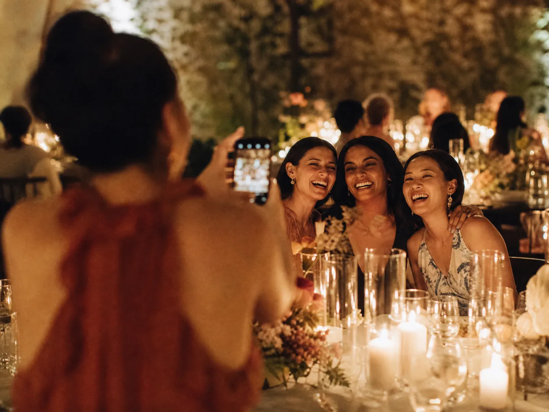 A guest holds up her phone to photograph friends laughing across a candlelit table.