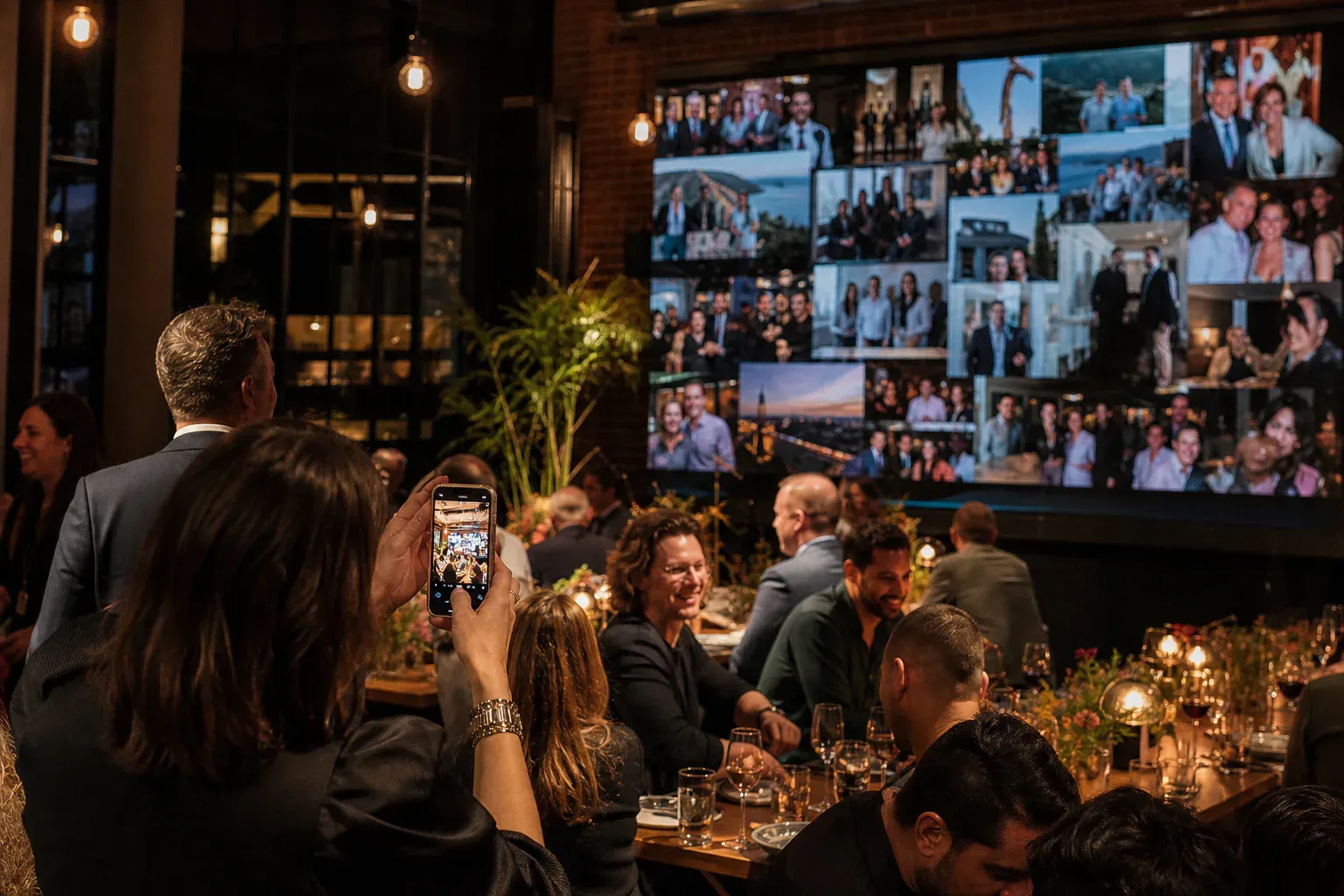 Guests taking candid photographs at a reception while a large screen displays a live collage from the room.