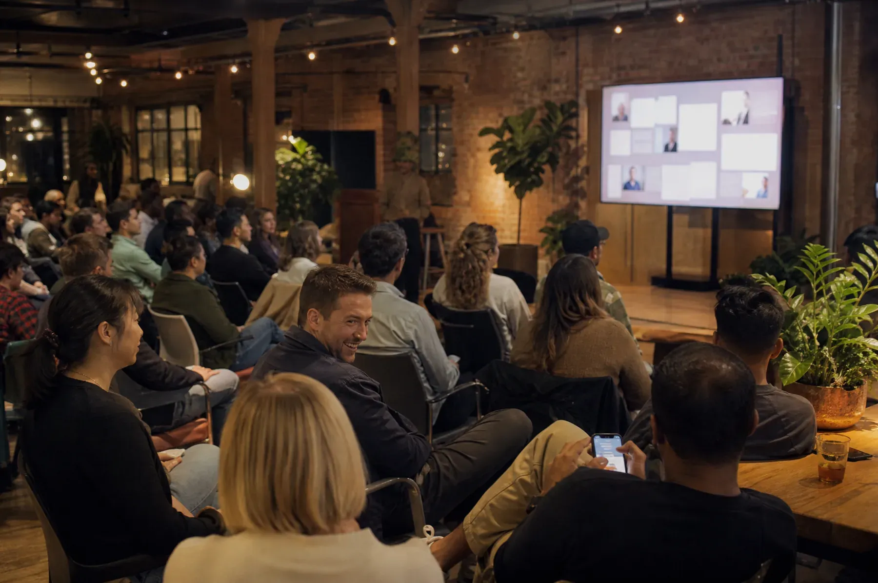 A company town hall in a converted warehouse space where a large screen beside the stage shows employee messages while colleagues watch from rows of seats.