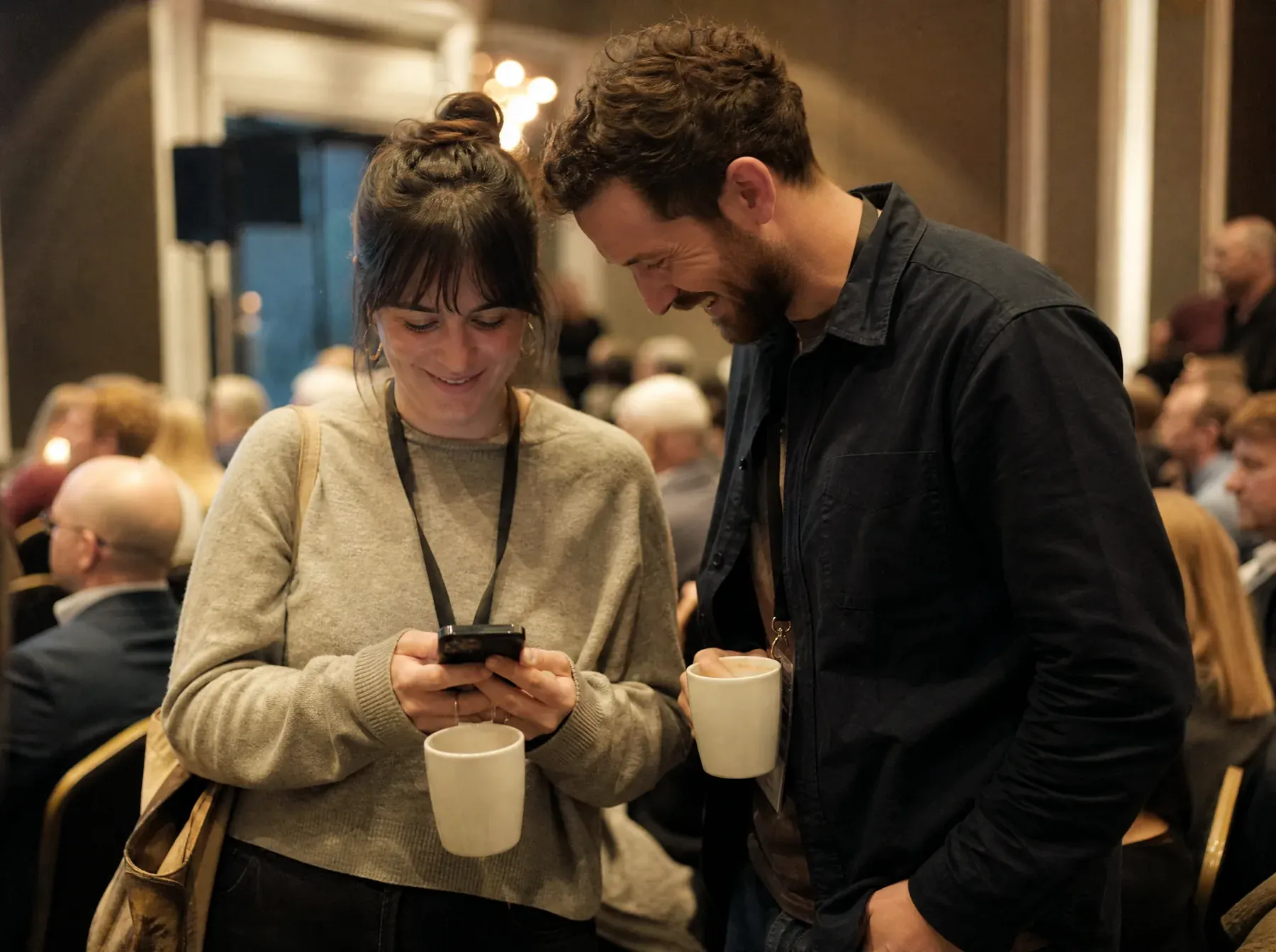 Two colleagues at the back of an internal event, one typing on her phone while the other reads over her shoulder and laughs.