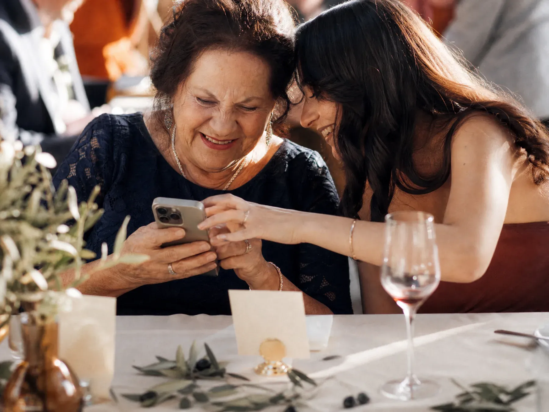 An older wedding guest types a message on her phone at a candlelit table while a younger relative leans in to help.